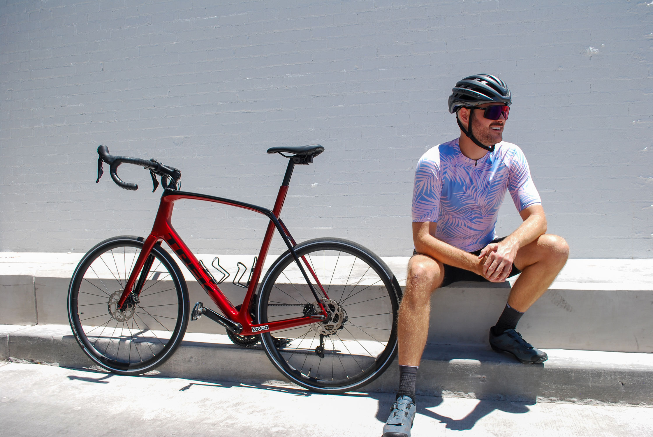 A rider in the Tropical Paradise jersey, sitting on a kerb beside a red road bike